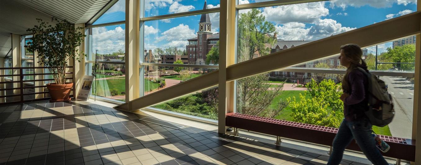 student wearing backback walking through halway with floor to ceiling windows. A beautiful sunny green campus outside